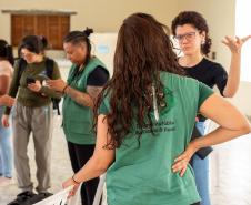 Equipe da Defensoria realiza atendimentos em localidade no Litoral do Paraná. Imagem mostra Defensora Pública de costas usando colete da instituição conversando com uma mulher. Ao fundo é possível ver mais servidores falando com pessoas em atendimento. 