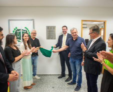 Um grupo de sete pessoas, entre homens e mulheres, sorri e bate palmas enquanto duas delas puxam um tecido verde para inaugurar uma placa prateada fixada na parede branca de um corredor.