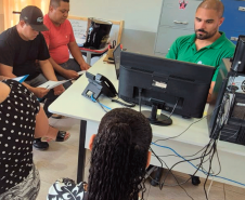 Uma sala com um funcionário de camisa verde atendendo em um computador. À frente dele, um grupo de pessoas aguarda sentado.