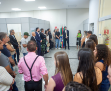 Uma mulher de blusa verde fala ao microfone em um pequeno púlpito, de frente para uma plateia que a assiste de pé em uma sala ampla. Atrás dela, junto a bandeiras oficiais, estão outras autoridades.