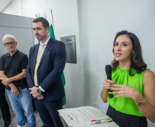 Em um plano mais aproximado, a mulher de blusa verde discursa ao microfone com expressão atenta, apoiada em um púlpito com papéis. Logo atrás dela, três homens aguardam em pé, escutando-a com seriedade.