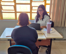 Próximo a uma grande janela com esquadrias amarelas, uma mulher de óculos e camisa branca digita em um notebook. Sentada de costas em uma cadeira azul, uma mulher de roupa escura recebe o atendimento.