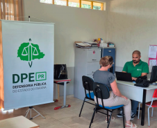 Visão mais ampla da sala com um banner vertical da DPE-PR em destaque à esquerda. Ao fundo, o funcionário de verde usa o notebook enquanto atende uma mulher de blusa cinza sentada à sua frente.