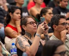 Imagem mostra detalhe do público presente. Em primeiro plano uma mulher com tatuagens registra o evento com celular.