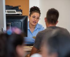 Imagem mostra uma servidora da DPE-PR realizando atendimento a um cidadão. Ele aparece de costas. Em primeiro plano desfocado aparecem cabeças de pessoas na fila de espera.