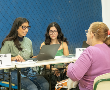 Imagem mostra uma mulher com cabelos presos de costas recebendo atendimento de duas servidoras da Defensoria Pública do Paraná.