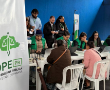 Em um espaço com parede azul, profissionais com coletes verdes usam notebooks em mesas brancas para atender cidadãos sentados à sua frente. À esquerda, destaca-se um banner vertical da Defensoria Pública do Paraná.