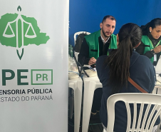 Close-up de um atendimento onde um servidor de colete verde conversa com uma mulher sentada de costas. Ao lado dele, outra profissional manuseia o celular, com um banner da DPE-PR em grande destaque no lado esquerdo.
