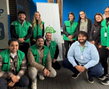 Dez profissionais da equipe da DPE-PR posam sorridentes para uma foto em grupo. A maioria veste coletes verdes da instituição, dividindo-se entre pessoas em pé e agachadas ao redor de um banner institucional.