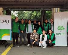 Uma equipe de doze pessoas posa para foto em um espaço público com piso tátil amarelo no chão. A equipe, com vários membros usando coletes verdes, está posicionada entre os banners da Defensoria Pública da União (DPU) e da DPE-PR.
