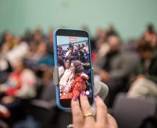 Imagem mostra close em celular mostrando o auditório cheio ao fundo.