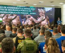 Imagem mostra o salão de atos do Palácio Iguaçu lotado com oficiais das forças de segurança. Ao fundo letreiro sobre o lançamento do NUSEG e o DPG Matheus Munhoz discursando.  