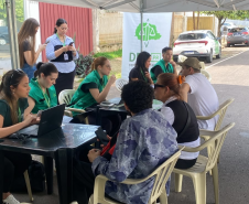 Em uma tenda de atendimento na rua a equipe da Defensoria Pública conversa e orienta as pessoas sentadas à frente das mesas. Ao fundo, destaca-se um banner branco e verde da DPE-PR.