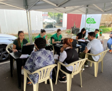 Atendimento ao ar livre sob uma tenda branca. Funcionárias de colete verde da DPE-PR utilizam notebooks em mesas para atender cidadãos que estão sentados em cadeiras de plástico branco.