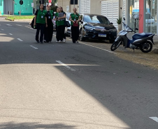 Quatro mulheres vestindo coletes verdes da Defensoria Pública caminham juntas e sorridentes por uma rua asfaltada. Elas seguram pastas e passam ao lado de veículos estacionados em uma área urbana.