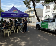 Uma tenda azul com a inscrição "Centro da Juventude" montada na rua abriga pessoas em atendimento. Estacionada logo ao lado, sob a sombra de uma árvore, está uma caminhonete branca com a logo verde da DPE-PR.