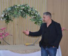 Um homem de blazer escuro, camisa preta e calça jeans gesticula com a mão direita. Ele está posicionado em frente a um arco decorativo adornado com flores brancas e folhagens, ladeado por arranjos de flores rosas.