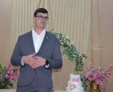 Um homem de terno cinza, camisa branca e óculos está de pé com as mãos cruzadas à frente, olhando levemente para o lado. Próximo a ele há um bolo branco de três andares enfeitado com flores rosas, com o arco floral ao fundo.