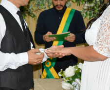 Detalhe do momento exato da troca de alianças. O noivo, de colete preto e camisa branca, coloca a aliança no dedo da noiva de vestido rendado, enquanto o celebrante com faixa verde e amarela observa desfocado ao fundo.