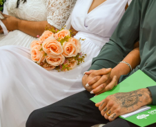 Close no colo de um casal sentado. A noiva, de vestido branco, segura um buquê de rosas pêssego, enquanto o companheiro ao lado apoia as mãos, uma delas tatuada e com aliança, sobre uma pasta verde da DPE-PR.