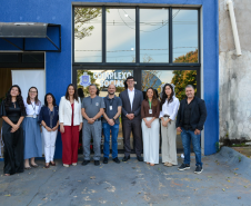 Um grupo de dez pessoas posa sorridente para uma fotografia em frente à fachada azul e às portas de vidro de um prédio identificado como "COMPLEXO SOCIAL". A equipe veste trajes sociais casuais, e alguns usam crachás de identificação no pescoço.