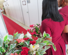 Um casal, com a mulher vestindo um vestido vermelho, caminha por um corredor coberto por um tapete vermelho. Em primeiro plano e ligeiramente desfocados, arranjos de flores brancas, rosas e vermelhas enfeitam a lateral do caminho.