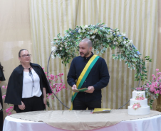 Um celebrante com uma faixa verde e amarela no peito está posicionado atrás de uma mesa com um pequeno bolo de casamento enfeitado com flores. Ao lado dele, uma mulher de blusa preta e branca acompanha a cerimônia em frente a um arco floral.