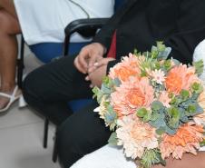 Close nas mãos de uma noiva segurando um buquê de flores em tons de pêssego, alaranjado e folhagens verdes. Ela veste um vestido branco com detalhes em renda e está sentada ao lado de um homem de terno preto e gravata vermelha.