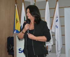 Uma mulher de cabelos escuros e conjunto preto segura um microfone enquanto fala. Logo atrás dela, estão posicionadas mastros com bandeiras oficiais e parte de um banner da Defensoria Pública.