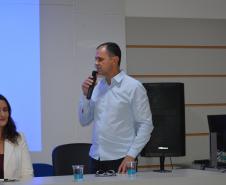 Um homem de camisa azul clara fala ao microfone em pé. Ao lado, uma mulher de blazer branco está sentada à mesa. Ao fundo, há uma tela de projeção exibindo uma luz azul e uma caixa de som preta.