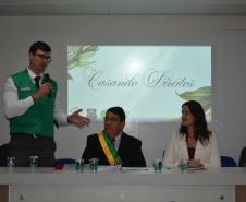 Um homem de colete verde fala ao microfone em pé ao lado de uma mesa com três pessoas sentadas. Atrás deles, uma projeção exibe a frase "Casando Direitos". Na mesa, destaca-se um homem com uma faixa verde e amarela no peito.