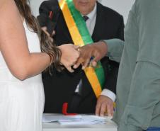 Close de um casal trocando alianças em uma cerimônia. A noiva veste branco e o noivo veste uma camisa verde. Ao fundo, o celebrante que segura o microfone usa uma faixa verde e amarela, e uma caixinha vermelha de anel repousa sobre a mesa.