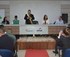 Visão ampla de um auditório onde um celebrante com faixa verde e amarela fala ao microfone em uma mesa diretiva com as logos do Fecomércio PR e Senac. À esquerda, há um banner da DPE-PR. O público assiste sentado, com pessoas fotografando a cena.