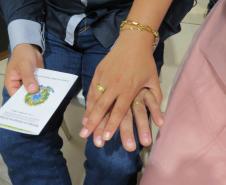 Foco nas mãos cruzadas de um casal em que ambos exibem alianças douradas. Uma das pessoas veste calça jeans e segura um documento da "República Federativa do Brasil", enquanto parte de um vestido rosa brilhante aparece à direita.