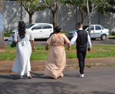 Visão de costas de três pessoas caminhando de mãos dadas por uma rua asfaltada e arborizada com carros estacionados. Da esquerda para a direita, caminham uma noiva de vestido branco, uma mulher de vestido longo bege e um noivo de colete preto e camisa branca.