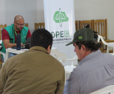 Um servidor de óculos e colete verde atende em um notebook numa mesa branca. À sua frente, dois homens aguardam sentados de costas. Ao fundo, há um banner vertical da DPE-PR.