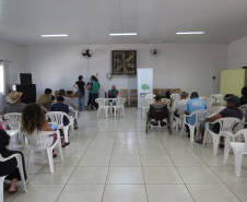Visão ampla de um salão com diversas pessoas sentadas em cadeiras brancas voltadas para o fundo. Lá, servidores da DPE-PR estão próximos a mesas posicionadas ao lado de um banner da instituição.