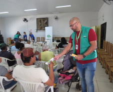 Um funcionário em pé, de óculos e colete verde, entrega um panfleto a um homem sentado de costas. Ao lado do servidor há um carrinho com um bebê dormindo, e outras pessoas aguardam ao fundo do salão.