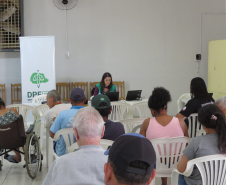 Uma mulher de colete verde trabalha em um notebook ao fundo da sala, ao lado de um banner da DPE-PR. À frente dela, diversas pessoas aguardam sentadas de costas, incluindo uma pessoa em cadeira de rodas.