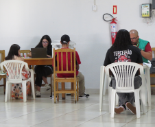 Duas mesas de atendimento simultâneo. À esquerda, uma mulher no notebook atende duas pessoas. À direita, um homem de colete verde atende um jovem vestido com uma camisa preta com a palavra "TERCEIRÃO" nas costas.