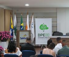 Um auditório com o público sentado de costas em cadeiras azuis, voltado para um palco. No palco, há um púlpito com a logo do Senac acompanhado por bandeiras oficiais, e à direita destaca-se um banner da Defensoria Pública (DPE-PR).