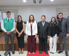 Seis pessoas posam lado a lado em um palco de piso de madeira com um tapete vermelho no centro. O grupo é formado por homens e mulheres em trajes sociais, incluindo, à extrema esquerda, um homem vestindo o colete verde da DPE-PR.