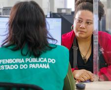 Imagem mostra servidor de costas com colete verde onde se lê Defensoria Pública do Parané e ao fundo uma mulher assistida durante Mutirão Meu Pai Tem Nome.
