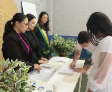 Um homem de camiseta branca assina um documento sobre uma mesa, ao lado de uma mulher de vestido branco. Atrás da mesa, três mulheres observam atentamente, sendo que uma delas usa uma faixa transversal com as cores da bandeira do Brasil.