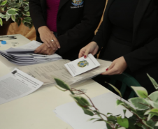 Detalhe de uma mesa com vários documentos impressos. Duas mulheres de terno preto manuseiam os papéis, e uma delas segura um livreto com o brasão da República do Brasil na capa. À esquerda, vê-se a mão de uma pessoa assinando um papel.
