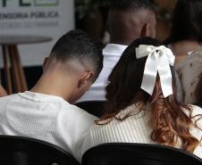 Visão traseira de um casal sentado em meio a uma plateia. O homem tem cabelo escuro com corte em degradê, e a mulher tem cabelos longos e ondulados, enfeitados na parte de trás com um grande laço branco.