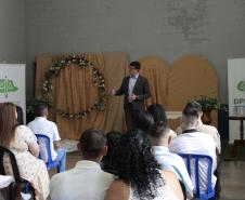 Um homem de terno escuro discursa em pé para o público acomodado em cadeiras de plástico azul e preta. Ele está em frente à decoração dourada com um arco de flores, ladeado por um banner da DPE-PR à direita.