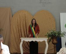 Uma mulher com blazer vermelho e a faixa transversal verde e amarela está em pé atrás da mesa branca, falando aos presentes. O cenário mantém os painéis dourados ao fundo e o banner da DPE-PR à direita.