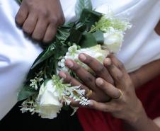 Detalhe em close de mãos negras entrelaçadas sobre um buquê de rosas brancas, ambas exibindo alianças douradas. Uma das pessoas veste um traje branco (vestido), e a outra usa uma blusa vermelha.