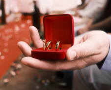 A mão de um homem, segura uma caixinha vermelha com três alianças de casamento. 